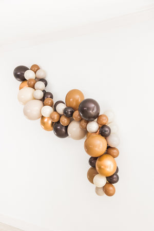 Side view of a balloon garland with the colors brown, mocha, tan, double stuffed tan, and copper hangs on a white wall.