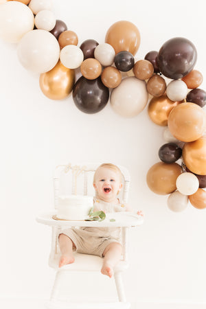 Baby in a white high chair smiling with a balloon garland with the colors brown, mocha, tan, double stuffed tan, and copper above him.