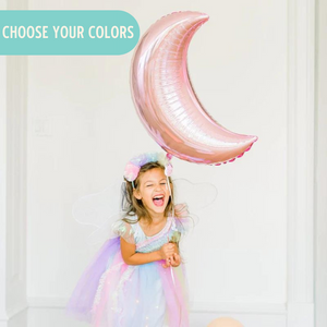 Little girl wearing a fairy costume is smiling and holding a pink crescent moon jumbo balloon.