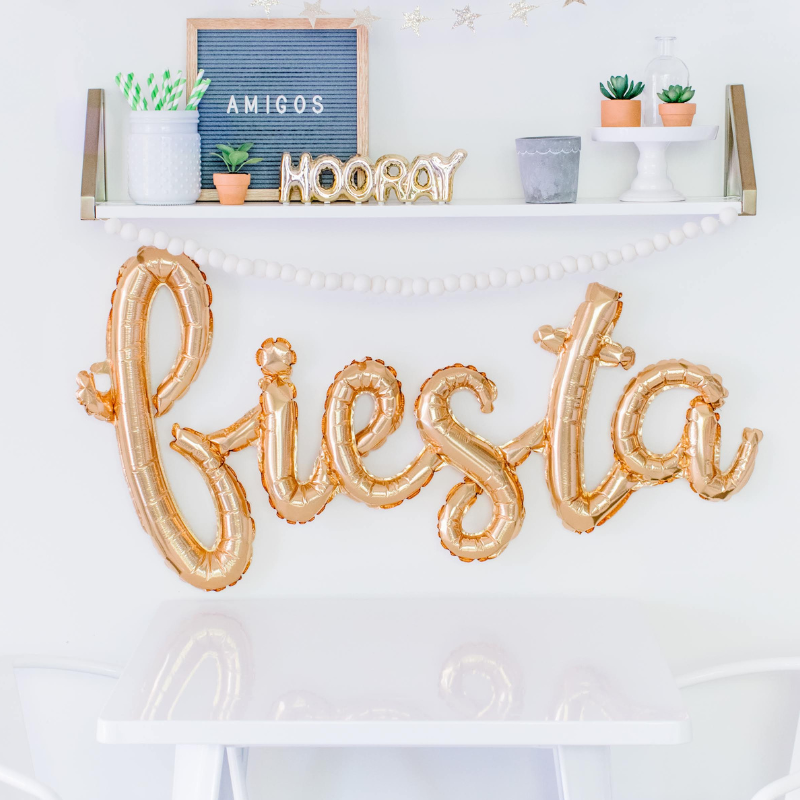 A gold cursive fiesta script balloon is strung on a wall underneath some white floating shelves above a small white kids table and chairs.