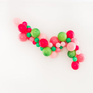 A fun summery watermelon themed garland sits on a white wall. Made up of pink, rose, wildberry, spring green, and green marble latex balloons in 5 inch and 11 inch sizes.