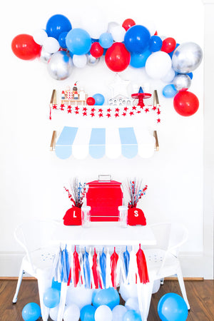 A perfectly patriotic themed balloon garland displayed on a white wall with a themed tassel garland hanging underneath. Balloons are a mix of red, blue, light blue, chrome silver, and white. Tassels are in matching red, white, blue, and silver.