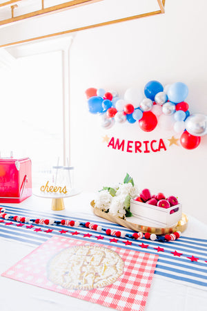 A 4th of July themed balloon garland displayed on a white wall. Balloons featured in the garland are red, blue, light blue, chrome silver, and white.