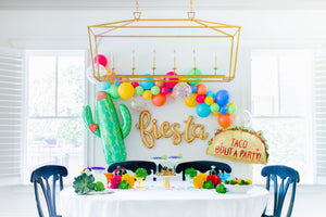 A gold cursive fiesta script balloon is strung on a wall surrounded by a colorful fiesta style balloon garland, a giant cactus balloon, and a taco balloon overlooking a fiesta tablescape.