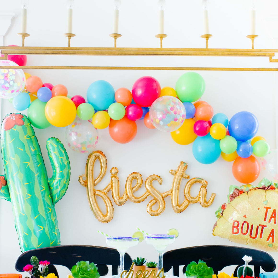 A gold cursive fiesta script balloon is strung on a wall surrounded by a colorful fiesta style balloon garland, a giant cactus balloon, and a taco balloon.