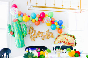 Close up of a gold cursive fiesta script balloon strung on a wall surrounded by a colorful fiesta style balloon garland, a giant cactus balloon, and a taco balloon.