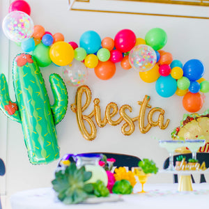 A gold cursive mylar fiesta script balloon is strung on a wall surrounded by a colorful fiesta style latex balloon garland, a giant mylar cactus balloon, and a taco mylar balloon.