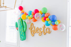 Side view of a gold cursive fiesta script balloon hanging from a white wall surrounded by a colorful fiesta style balloon garland and a giant cactus balloon.