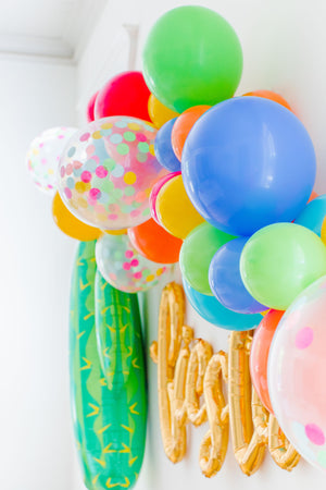 Side view of a gold cursive fiesta script balloon hanging from a white wall surrounded by a colorful fiesta style balloon garland and a giant cactus balloon.