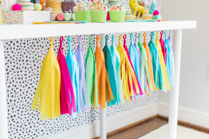 Side view of a tissue tassel garland in the colors yellow, pink, blue, purple, green, and orange hangs from a white table. Plastic zoo animals wearing mini party hats sit on top of a white table with cupcakes and macarons.