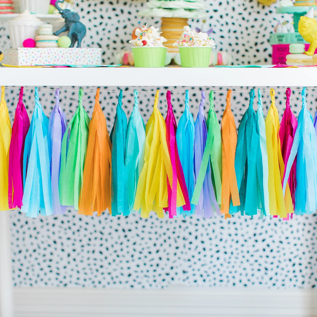 Tissue tassel garland in the colors yellow, pink, blue, purple, green, and orange hangs from a white table. Plastic zoo animals wearing mini party hats sit on top of a white table with cupcakes and macarons.