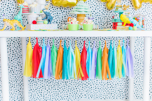 Tissue tassel garland in the colors yellow, pink, blue, purple, green, and orange hangs from a white table. Plastic zoo animals wearing mini party hats sit on top of a white table with cupcakes and macarons.