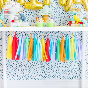 Tissue tassel garland in the colors yellow, pink, blue, purple, green, and orange hangs from a white table. Plastic zoo animals wearing mini party hats sit on top of a white table with cupcakes and macarons.