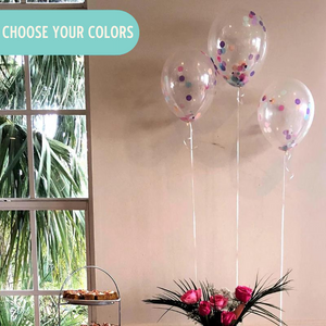 Three confetti balloons filled with rich purple, pink, and blue confetti pieces float above a table with a pink table cloth. The table is loaded with cakes and cookies sitting on various stands.