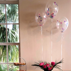 Three confetti balloons filled with rich purple, pink, and blue confetti pieces float above a table with a pink table cloth. The table is loaded with cakes and cookies sitting on various stands.
