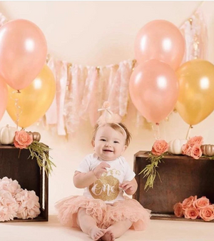 A little girl sitting on the ground with rose gold and gold balloons floating next to her.