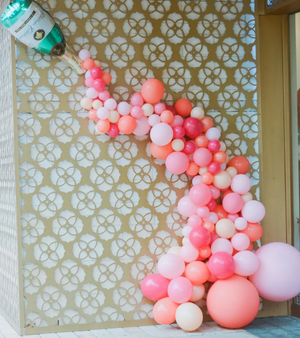 A tall wall with a champagne bottle balloon at the top left corner and pink, coral, and dark pink balloons in different sizes cascading out of the champagne bottle balloon.