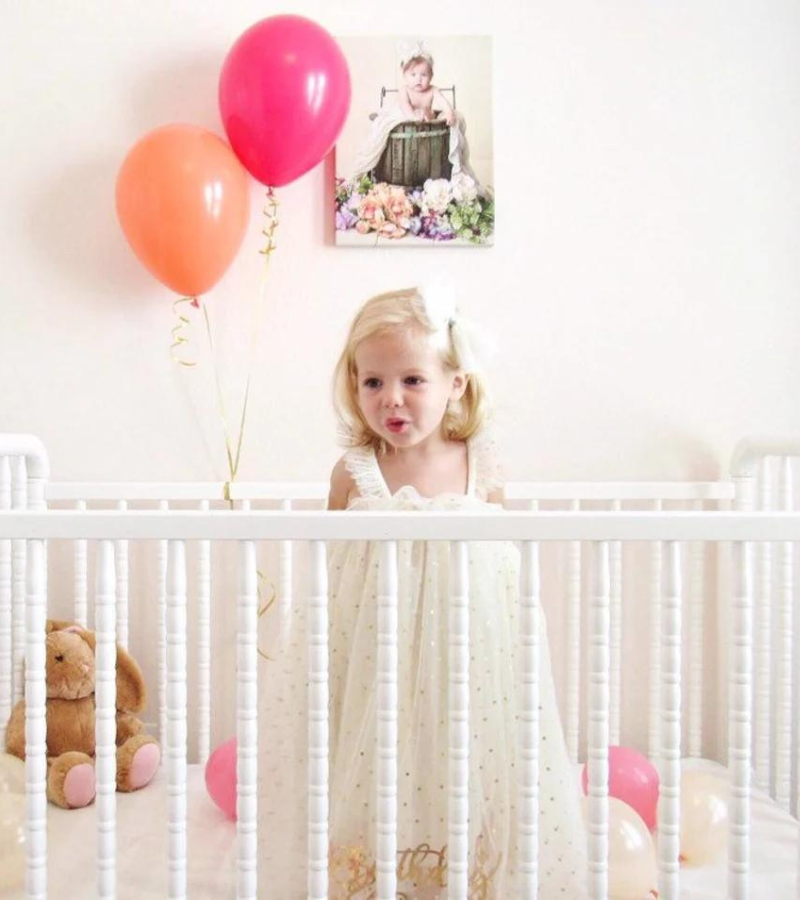 A little girl in a white crib holding a wildberry and orange balloon inflated with helium.