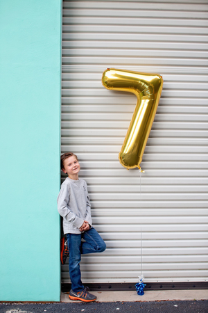 A boy smiling in front of a white colored garage door while holding a gold number seven balloon at his side.