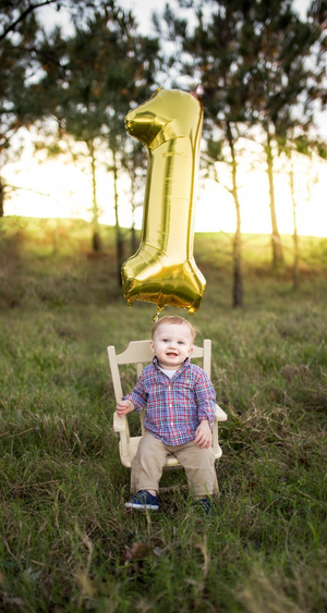 A little boy sitting on a small rocking chair in the grass with a gold number 1 balloon floating behind him.