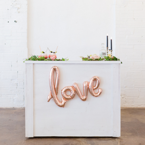 A cursive rose gold balloon spelling LOVE hung on a white bar.