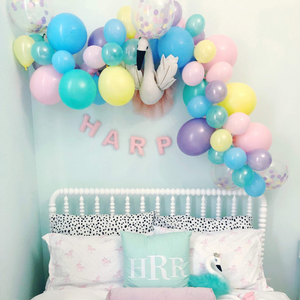 A pretty white metal framed bed sits below a pastel themed balloon garland strung across a mint colored wall. The balloon garland is made up of pink, pastel mint, pastel yellow, pastel blue, pastel lilac, and coordinating matching confetti balloons.
