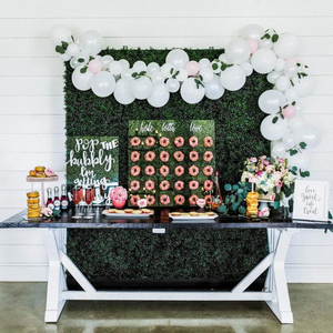 A donut assortment sits on top of a rustic table. There is a white balloon garland behind it hanging from a photo backdrop.