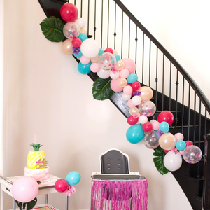 A balloon garland is displayed on a staircase railway. Below is a high chair and a pineapple theme cake.