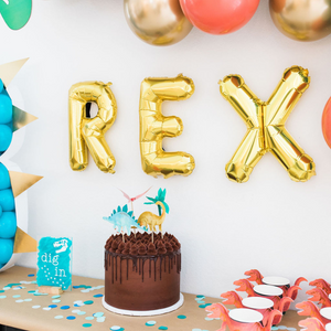 A long entry way table has blue, aqua, and teal tissue confetti spread around it with a brown cake and dinosaur themed cups sitting on top.