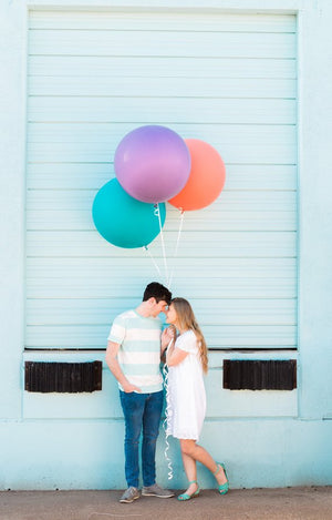 Young couple standing in front of a blue wall holding three jumbo 36 inch purple, coral, and lilac balloons.