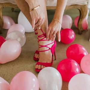 A view of a woman tying a hot pink high heel with various pink and white 5 inch balloons inflated and scattered all around the ground by her.