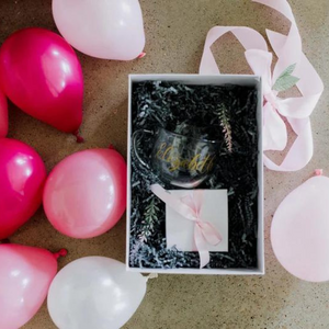 An arial shot of a pretty glass cup inside of a box with various pink colored 5 inch balloons inflated and scattered all around the box.