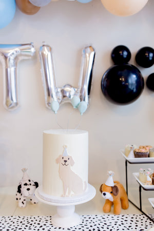 Jumbo silver letter balloons one with a T and the other with a W continued by black balloons making a pawprint, together they spell TWO. There is a white table below with a white cake and stuffed animal dogs wearing party hats.