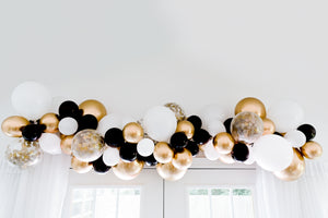 Black, white, gold, and clear confetti balloon garland hangs over a white door and drapes.