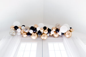 Black, white, gold, and clear confetti balloon garland hangs over a white door and drapes.