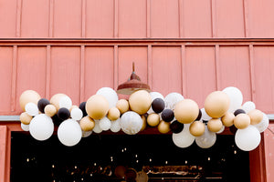 Black, white, gold, and clear confetti balloon garland hangs over a rustic barn building.