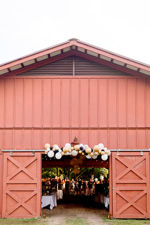Black, white, gold, and clear confetti balloon garland hangs over barn building.