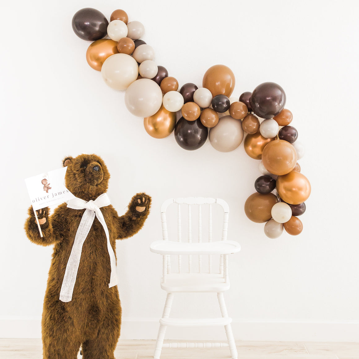 A life size bear is holding a white banner and standing next to a white chair. Above the chair is a balloon garland with the colors brown, mocha, tan, double stuffed tan, and copper. 