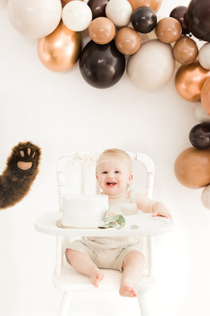 Balloon garland with the colors brown, mocha, tan, double stuffed tan, and copper hangs on a white wall above a little baby in a white high chair.