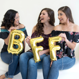 Three girls sit on a white cloth bench laughing at each other while each holding a gold mylar letter balloon. One holding a letter B, and the other two each holding a letter F.