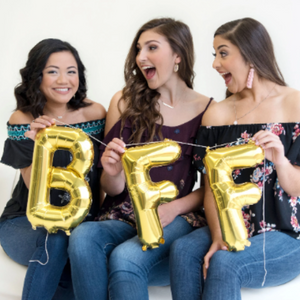 Three girls sit on a white cloth bench laughing at each other while each holding a gold mylar letter balloon. One holding a letter B, and the other two each holding a letter F.