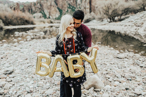 Man hugging a pregnant woman who is holding gold balloon letters that spell BABY.