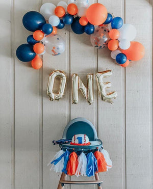 Little boys baseball theme one year old birthday party with silver 16 inch balloons spelling ONE and balloon arch garland with the colors blue, orange, and white.
