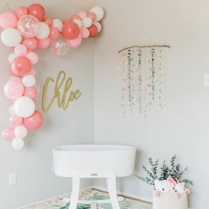 The same balloon garland sits on the corner wall of a white room with a pretty floral display and a gold wooden cutout of a child name underneath.