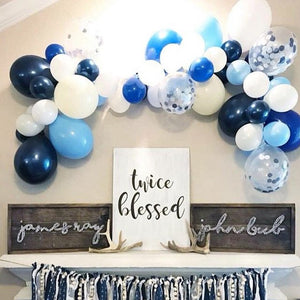 A balloon garland with different shades of blue, white, and clear balloons hangs from a wall above a party decorated table.