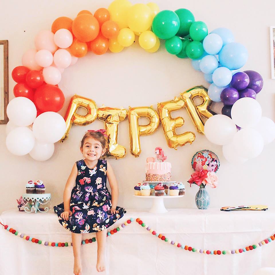 A little girl is smiling and sitting on top of a white party table. On the wall there is a rainbow balloon garland and jumbo gold letters spelling PIPER.