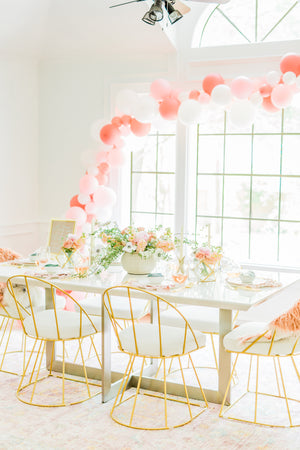 A big bright room with a white table full of flower bouquets has a soft balloon garland draped across the top of a bright white framed window. The balloons used in the garland are white, pink, mauve, and pink and blush confetti filled balloons.