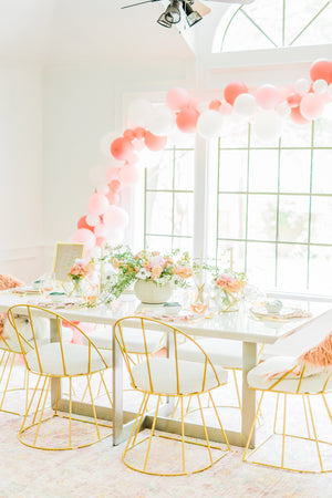 A big bright room with a white table full of flower bouquets has a soft balloon garland draped across the top of a bright white framed window. The balloons used in the garland are white, pink, mauve, and pink and blush confetti filled balloons.