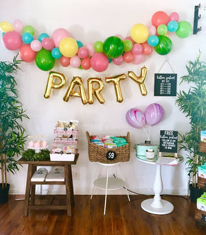 Giant gold letter balloons spelling PARTY hang on a white and a colorful balloon garland hangs above it.