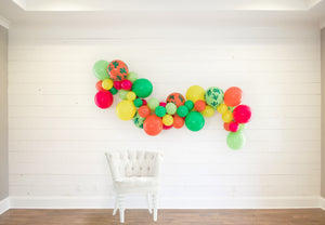 The balloon garland is draped on a white wood wall with a white clothe chair sitting in front to help show size and dimensions.